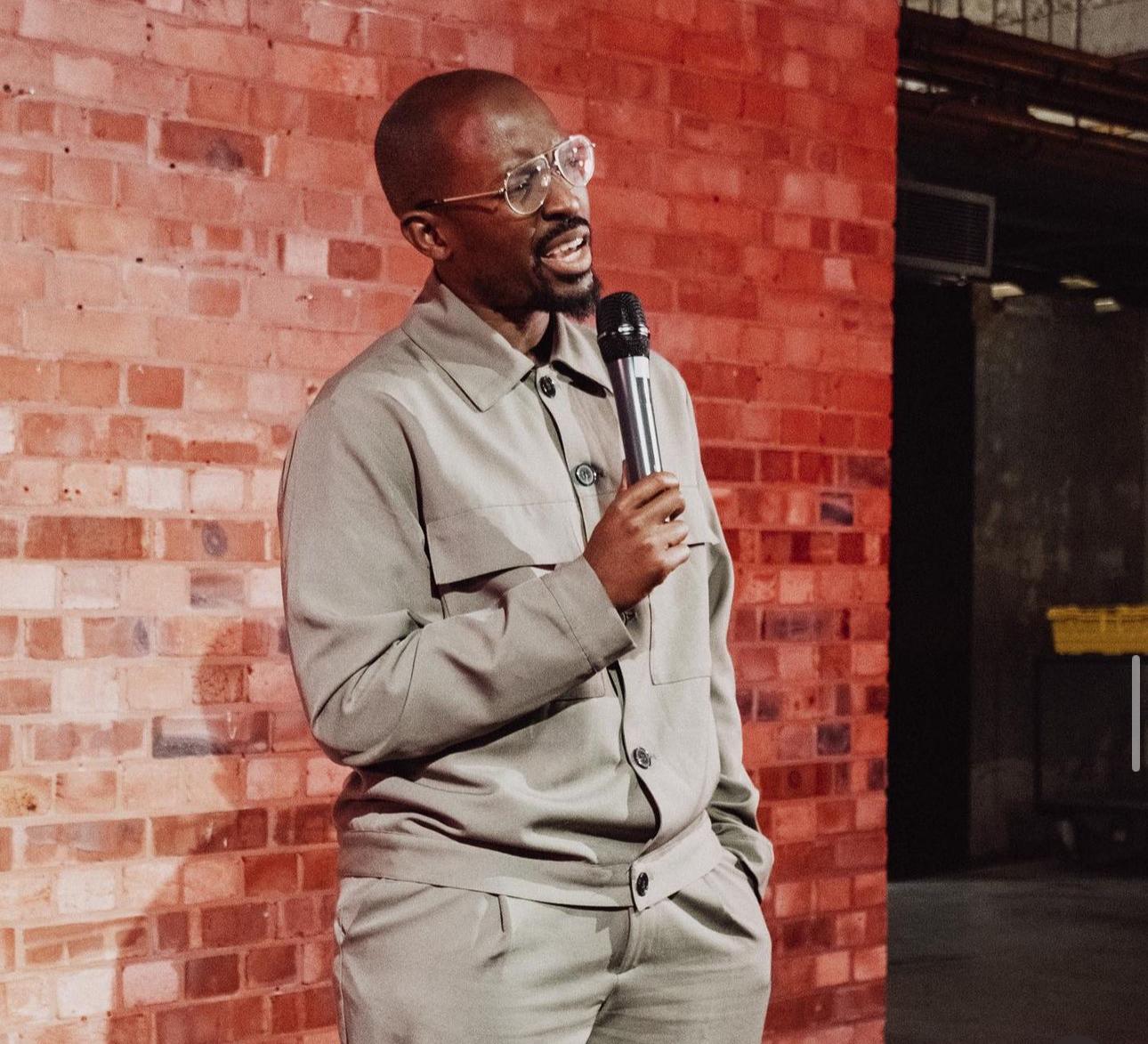 Person in beige outfit speaking into a microphone against a brick wall background.